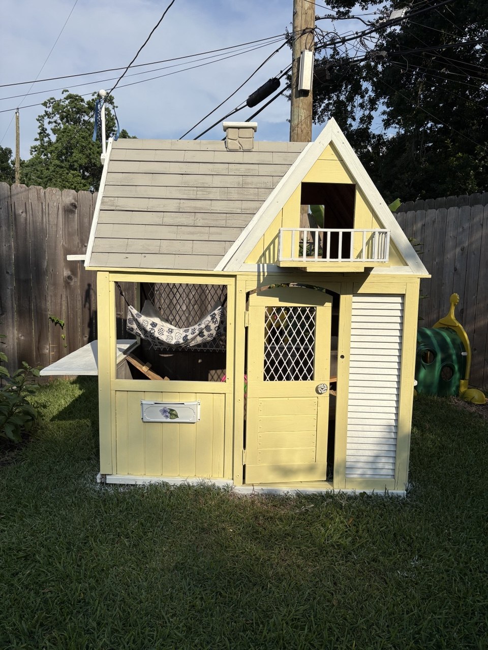 A two-story cat cottage painted pale yellow with white trim, a shingled roof, and a small balcony, standing on a lawn by a wooden fence